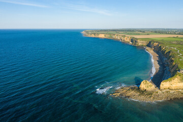 Obraz premium Sweeping aerial view of deep blue ocean meeting rugged cliffs and rocky shoreline at Pointe du Hoc, Normandy, with open farmland stretching beyond under a bright sky.