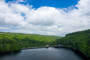 A long dam stretches across a dark, calm lake surrounded by dense green forest and rolling hills beneath a sky filled with large, billowing clouds at Lac de Chaumecon.