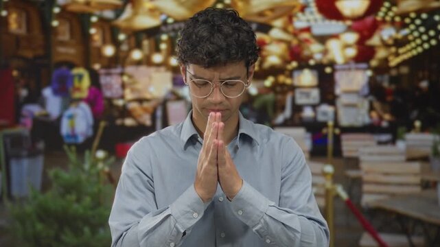 Man wearing glasses with clasped hands in prayer on street market under hanging lights; serenity.