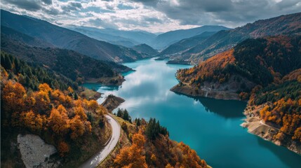 Obraz premium Aerial view of a vibrant lake surrounded by hills in autumnal colors, a winding road adds to the scenic view.