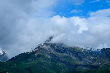 Dramatic clouds partially obscure a steep, rocky mountain with patches of snow and lush green slopes under a vibrant blue sky in the French Alps.