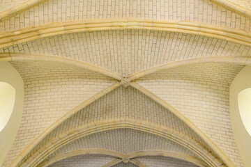 Detailed view of a pale stone vaulted ceiling featuring intersecting ribs and textured brickwork, illuminated by soft natural light. The architectural lines create a sense of symmetry and historic