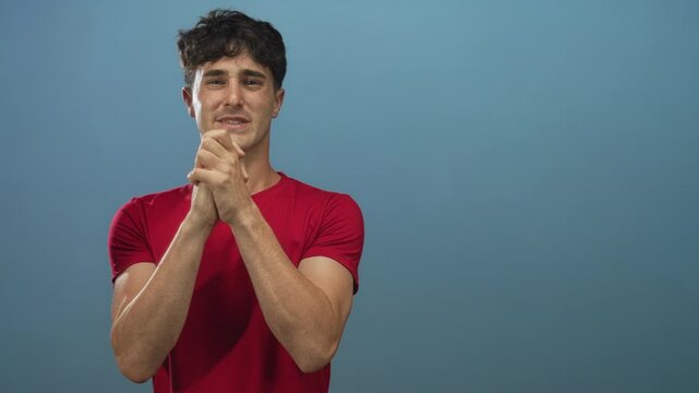 Young hispanic man with clasped hands and pleading facial expression in blue studio; pleading sincere hope.