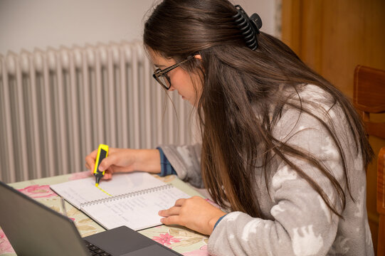 Teenage girl highlighting notebook with yellow marker while studying at home near laptop, wearing glasses and cozy pajamas