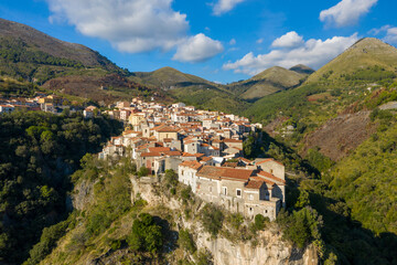 Vivid aerial view of Tortora, Italy, where rustic stone homes with red-tiled roofs cluster dramatically along the edge of a steep cliff, surrounded by rolling green hills under a bright blue sky with