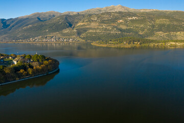 Fototapeta premium Aerial view of a tree-lined peninsula extending into Lake Ioannina, with tranquil water reflecting the rugged slopes of the Pindus mountains. The scene features clear daylight, natural textures, and a