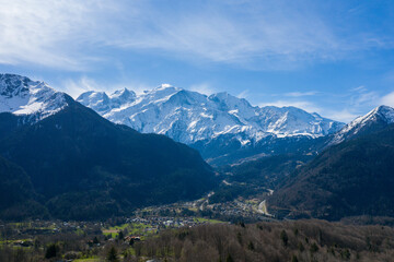 Fototapeta premium Wide view of the snow-covered Mont Blanc massif rising above a green alpine valley dotted with villages and surrounded by forested slopes under a bright blue sky.