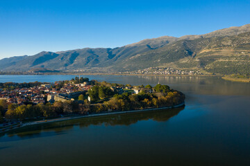 Fototapeta premium A wide aerial perspective captures the historic old town of Ioannina surrounded by Lake Ioannina, with the Pindus mountains rising in the background. The scene features red-tiled rooftops, medieval