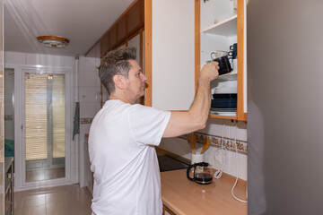 Side view horizontal of a real adult man in a home kitchen taking a mug from a cupboard while preparing coffee. Authentic everyday lifestyle scene in a real domestic environment. Concept of real life