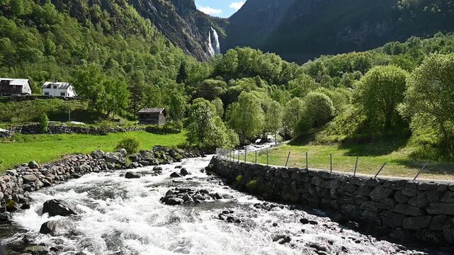 Calm river flows through lush green valley with waterfall and farm near Urnes, Norway.