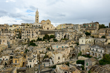 Obraz premium Expansive vista of Matera, Italy, reveals layered stone buildings, narrow stairways, and the iconic cathedral rising above the historic center under a cloudy sky. The weathered architecture and muted