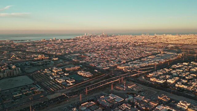 Expansive drone aerial view of a coastal city at sunrise, showcasing dense urban neighborhoods, skyline skyscrapers, and glowing golden light over the horizon&mdash;perfect for travel or architecture footag