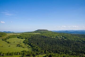 Fototapeta premium A solitary plateau rises above dense woodlands and rolling meadows under a clear blue sky in the Ardeche region, capturing the vastness and tranquility of the summer landscape.
