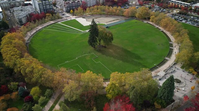 Aerial drone view of Bellevue Downtown Park featuring a large circular green lawn, central trees, and surrounding autumn foliage. Urban park landscape framed by walkways and modern buildings.