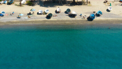 High-angle aerial view of a turquoise shoreline. Numerous swimmers are visible in the clear water, while the sandy beach is dotted with sunbathers and colorful umbrellas. It is a sunny summer morning.