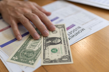 Hand placing US dollar cash on monthly budget worksheet on wooden desk. Concept of personal finance, budgeting, expense tracking, saving money, household accounting and financial planning.