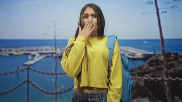 Woman student in yellow sweater with blue backpack covers mouth with hand at a harbor pier chain fence and boats; surprise concern.