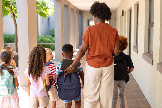 African American teacher wearing orange leading children along tiled hall with backpacks tablet