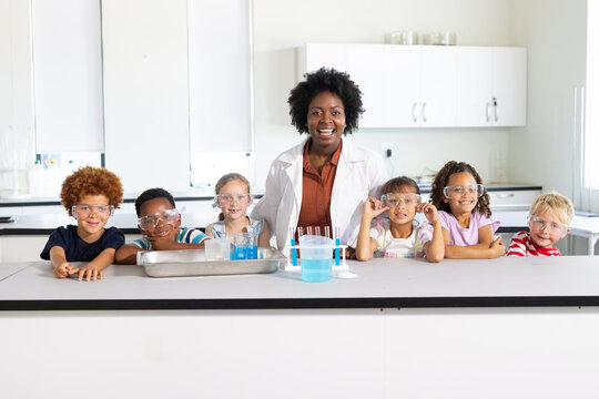 African American science teacher in lab coat guiding six children wearing safety goggles at bench
