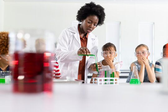 African American woman in white labcoat pouring liquid into test tube in lab, students in goggles