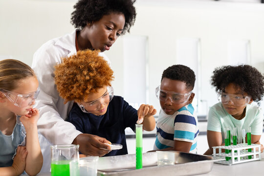 African American woman guiding kids handling green test tube at lab bench wearing lab coat, goggles