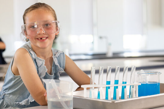 Preteen girl smiling wearing safety goggles and mixing blue liquids using test tubes at school lab