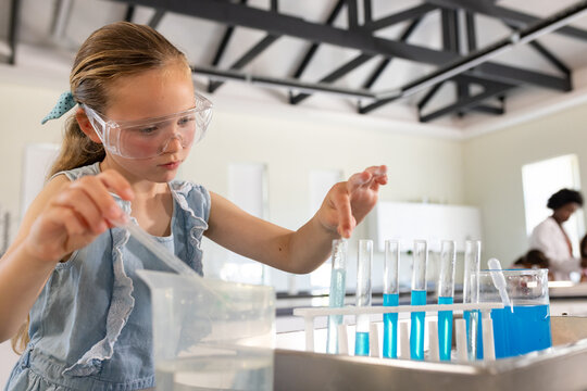 Diverse student and instructor in lab, girl child with goggles transferring liquid into test tubes