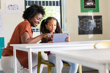 African American female teacher and youth female student using tablet in classroom with posters