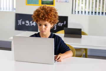 African American school-age boy working on silver laptop at classroom table near math poster