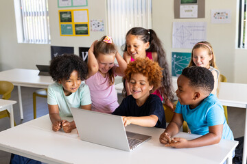 Diverse elementary children in tees leaning around silver laptop at school desk, pointing, laughing