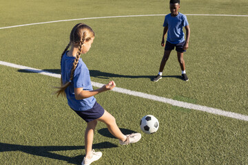 Diverse school-age children wearing blue shirts playing on turf pitch, girl controlling soccer ball
