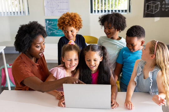 African American teacher with diverse children pointing at silver laptop at classroom table