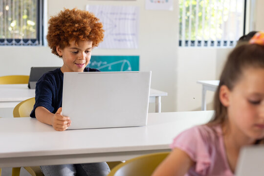 Diverse children with African American boy holding silver laptop working in classroom with posters
