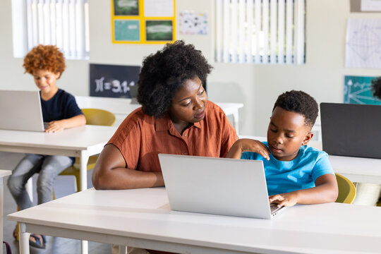 Female teacher in rust blouse leaning toward youth student guiding on silver laptop in class
