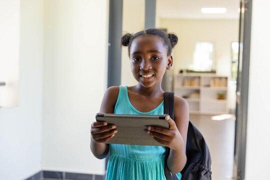 African youth standing holding tablet in hallway in turquoise dress and backpack with books behind - Powered by Adobe