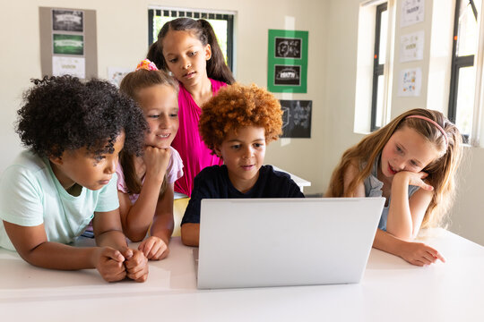 Diverse children leaning forward and watching silver laptop on white table in classroom
