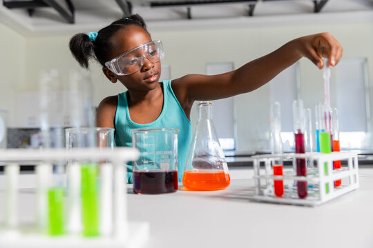 Female child wearing safety goggles and teal top reaching toward test tube rack at school lab