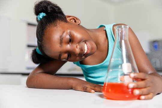 African youth in teal top leaning over table in lab, holding Erlenmeyer flask with orange liquid