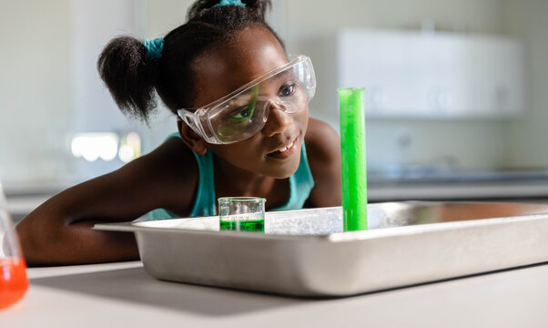 African teenage female leaning over lab wearing goggles observing green graduated cylinder, beaker
