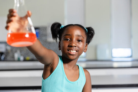 African American girl in turquoise top holding conical flask of orange liquid smiling in school lab
