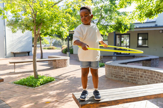 African American boy standing on wooden bench in sunlit brick courtyard holding yellow hula hoop