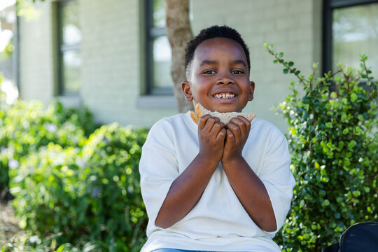 African American child sitting on low wall in yard, holding sandwich, backpack visible