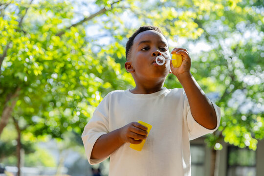 African male child standing in sunlit yard, blowing soap bubble with yellow wand and bottle