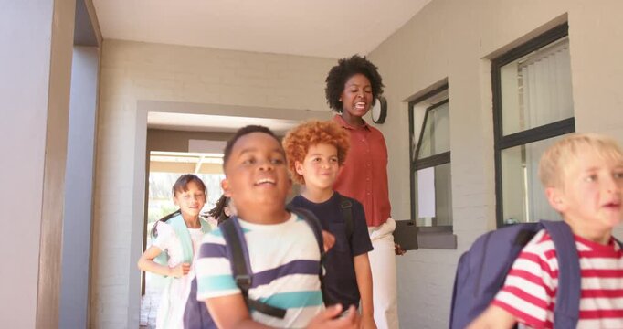 Diverse kids with backpacks exiting walkway toward yard, speeding with teacher holding tablet