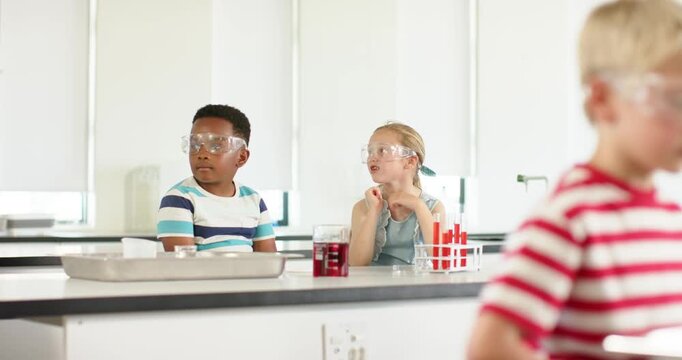 Diverse children at lab responding to teacher checking work, fixing goggles, watching red beaker
