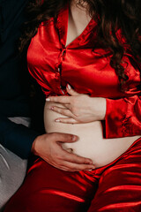 A pregnant girl in a red dress sits in the arms of her beloved man who is stroking her belly during a photo shoot