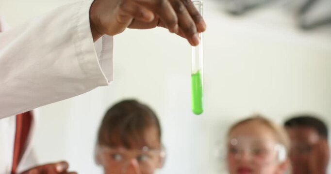 African American woman in lab coat leading demo, raising, tilting test tube green liquid for teens