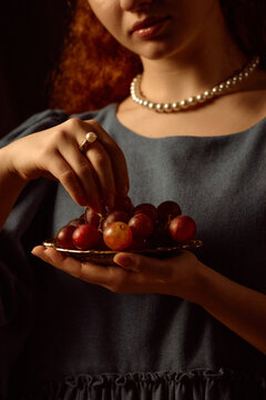 Young adult Caucasian woman holding plate of grapes, picking fruit with hand, wearing pearl necklace and ring, partial face visible, standing against dark background