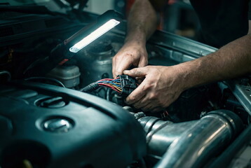 A focused mechanic's hands are illuminated by a work light, meticulously repairing complex electrical wiring in a car engine bay.