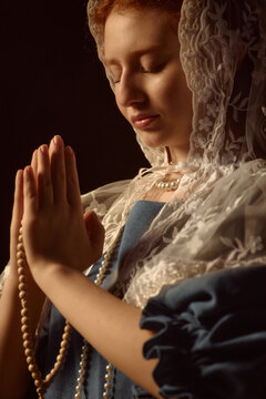 Caucasian young adult woman with red hair wearing lace head covering holding rosary beads and praying with closed eyes hands pressed together in gesture of devotion